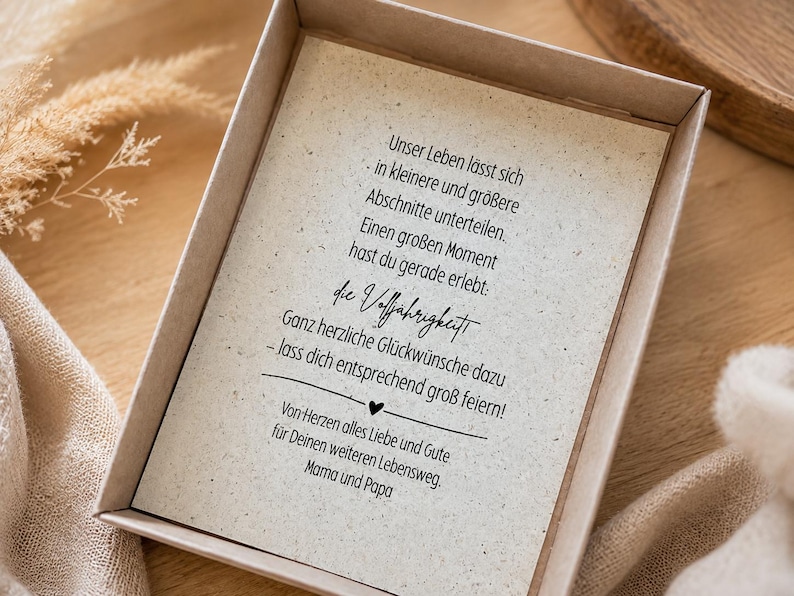 May include: A rectangular cardboard gift box containing a cream-coloured card with black printed German text. The text reads: Unser Leben l&auml;sst sich in kleinere und gr&ouml;&szlig;ere Abschnitte unterteilen. Einen gro&szlig;en Moment hast du gerade erlebt: die Vollj&auml;hrigkeit.