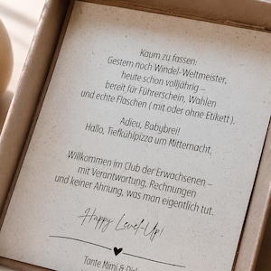 May include: A rectangular cardboard gift box containing a white card with black German text that reads: Kaum zu fassen: Gestern noch Windel-Weltmeister, heute schon vollj&auml;hrig &ndash; bereit f&uuml;r F&uuml;hrerschein, Wahlen und echte Flaschen. Happy Level-Up!