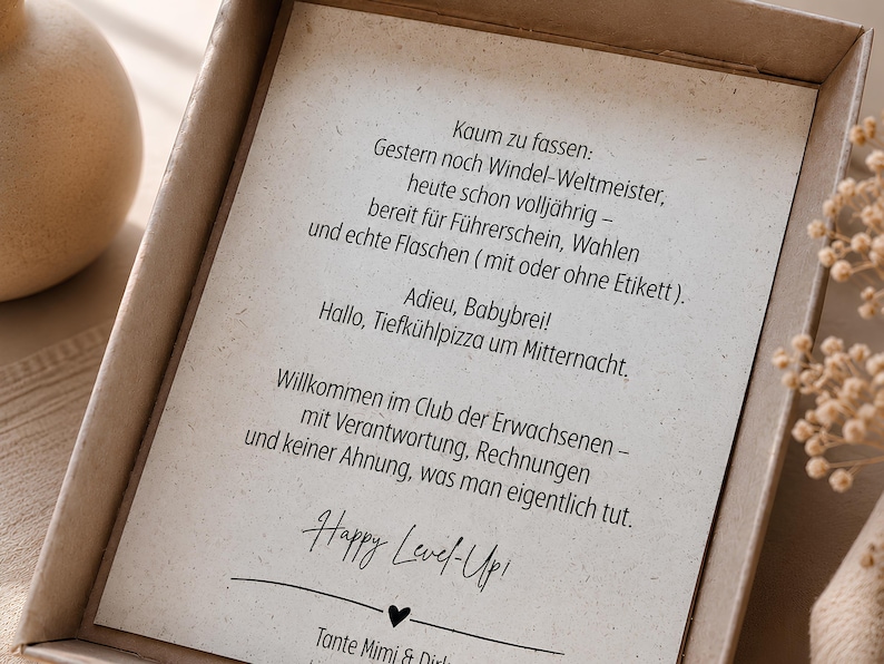May include: A rectangular cardboard gift box containing a white card with black German text that reads: Kaum zu fassen: Gestern noch Windel-Weltmeister, heute schon vollj&auml;hrig &ndash; bereit f&uuml;r F&uuml;hrerschein, Wahlen und echte Flaschen. Happy Level-Up!