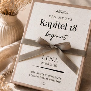May include: A rectangular gift box with a cream-coloured card featuring the German text "EIN NEUES Kapitel 18 beginnt", "LENA", "05.08.2026", and "DIE BESTEN MOMENTE LIEGEN NOCH VOR DIR." A taupe-coloured ribbon is tied in a bow across the centre of the box.