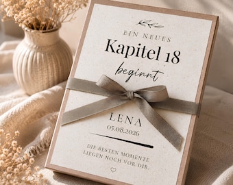 May include: A rectangular gift box with a cream-coloured card featuring the German text "EIN NEUES Kapitel 18 beginnt", "LENA", "05.08.2026", and "DIE BESTEN MOMENTE LIEGEN NOCH VOR DIR." A taupe-coloured ribbon is tied in a bow across the centre of the box.