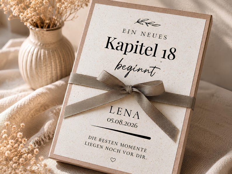 May include: A rectangular gift box with a cream-coloured card featuring the German text "EIN NEUES Kapitel 18 beginnt", "LENA", "05.08.2026", and "DIE BESTEN MOMENTE LIEGEN NOCH VOR DIR." A taupe-coloured ribbon is tied in a bow across the centre of the box.