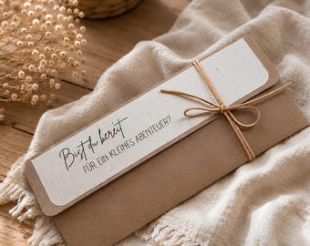 May include: A rectangular brown kraft paper envelope featuring a white paper insert with the black printed text "Bist du bereit FÜR EIN KLEINES ABENTEUER?" tied with a thin brown twine string in a bow across the front of the envelope.