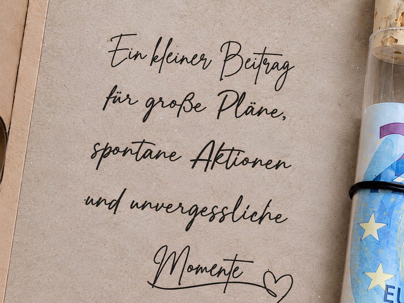 May include: A light brown card with black cursive text that reads: Ein kleiner Beitrag f&uuml;r gro&szlig;e Pl&auml;ne, spontane Aktionen und unvergessliche Momente, accompanied by a heart symbol. Next to it is a glass tube with a cork stopper containing a rolled banknote.