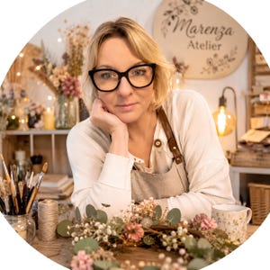 May include: A person with blonde hair and black-rimmed glasses wearing a beige linen apron over a white long-sleeved shirt. In front are paintbrushes in a glass jar, a spool of twine, a floral wreath, and a mug. A sign reads Marenza Atelier.