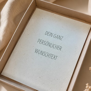 May include: A rectangular card with a speckled, off-white surface placed inside a light brown cardboard box. The card features black printed text that reads: DEIN GANZ PERS&Ouml;NLICHER WUNSCHTEXT. The card has a textured appearance with small dark flecks.