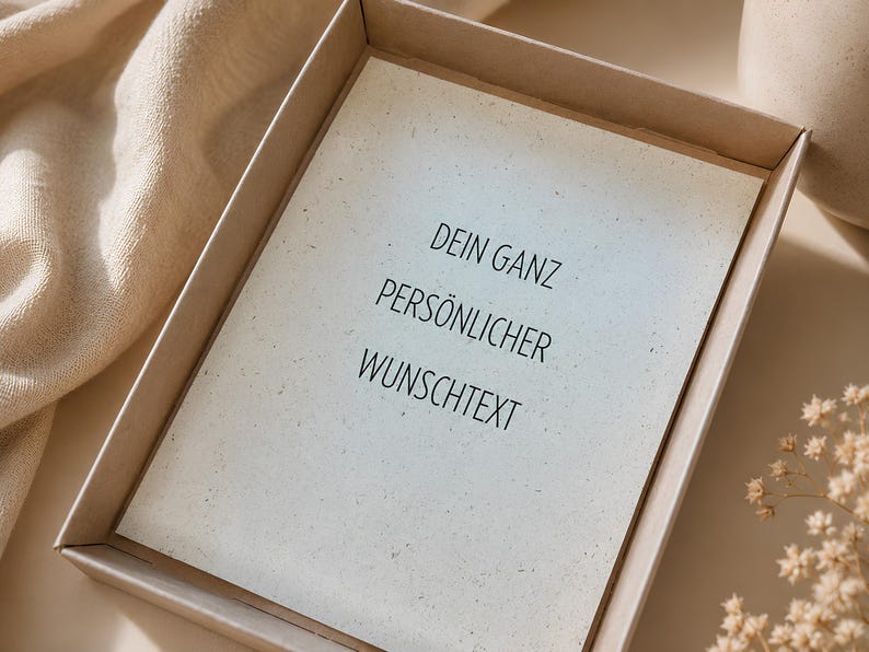 May include: A rectangular card with a speckled, off-white surface placed inside a light brown cardboard box. The card features black printed text that reads: DEIN GANZ PERS&Ouml;NLICHER WUNSCHTEXT. The card has a textured appearance with small dark flecks.