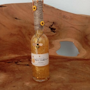 Peut inclure: Une bouteille en verre transparent remplie d'un liquide doré à paillettes, surmontée de coulures de cire beige et entourée de ficelle avec deux petits tournesols jaunes. Une breloque abeille pend au col, et une étiquette indique Elixir de Nectarea.