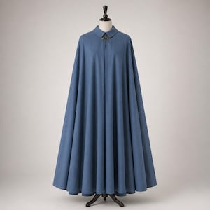 May include: A floor-length, solid blue cape displayed on a mannequin. The garment features a structured collar, a central front closure with a metallic clasp, and a full, pleated silhouette that drapes evenly from the shoulders to the hemline.