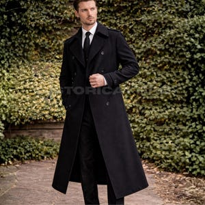 May include: A long, black double-breasted overcoat featuring a wide lapel, structured shoulders, and a row of dark buttons. The coat is worn over a white collared shirt and a black necktie, extending down to the lower calf with a clean, straight hem.