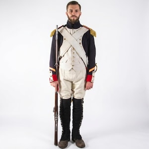 May include: A historical military uniform featuring a dark blue coat with gold epaulets, red cuffs, and white lapels. The ensemble includes a white vest, white breeches, and black gaiters. A long wooden musket is held vertically in one hand.