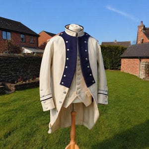 May include: A cream-colored long coat with a high collar and dark blue lapels, featuring silver-toned buttons down the front and on the cuffs. The coat is worn over a matching cream-colored vest, both displayed on a wooden mannequin stand.