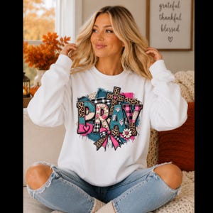 May include: A white long-sleeve sweatshirt featuring a graphic print of the word PRAY in a patchwork style. The letters incorporate leopard print, pink plaid, and floral patterns, accented by a cross, a bow, and a small flower design.