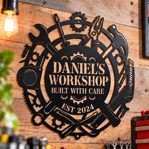 May include: A circular black metal wall sign featuring the text DANIEL'S WORKSHOP BUILT WITH CARE EST 2024. The border is composed of various tools including a wrench, screwdriver, pliers, hammer, and saw, interspersed with several gear shapes.