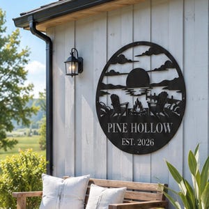May include: A circular black metal wall sign featuring a laser-cut landscape design of two chairs, a campfire, a lake, and a sun. The text PINE HOLLOW and EST. 2026 is displayed in the lower section of the circular frame, mounted on a white wall.