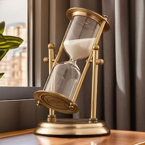 May include: A brass-coloured metal hourglass featuring a rotating frame and a clear glass bulb filled with white sand. The structure includes a circular base and top plate, with the glass component held securely within a pivoting support mechanism.