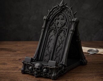 May include: A black phone stand featuring intricate gothic arch architecture with pointed details and vine-like carvings. The base is decorated with three sculpted skulls integrated into the frame, providing a stable support for a mobile device.