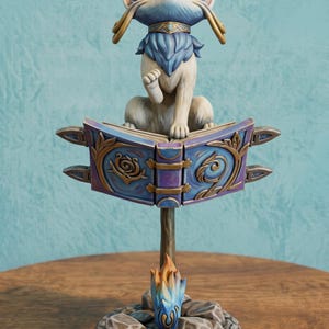 May include: A painted figurine of a cat-like creature with blue and cream fur sitting atop an open, purple, ornate book. The book is supported by a central post rising from a circular base featuring gray stones and a small, stylized blue and orange flame.