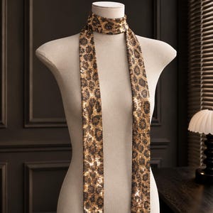 Puede incluir: Una bufanda larga y estrecha con un estampado de leopardo en dorado y negro cubierto de lentejuelas brillantes. La bufanda está envuelta alrededor del cuello de un maniquí, con dos extremos largos y planos que cuelgan verticalmente.