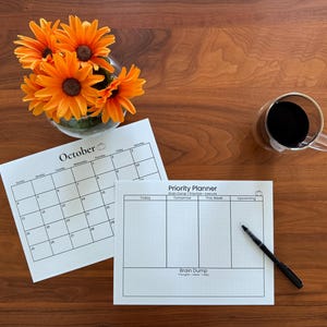 May include: A white October calendar and a white Priority Planner with columns for Today, Tomorrow, This Week, and Upcoming, featuring a Brain Dump section. A black pen rests on the planner, near a glass of dark liquid and orange flowers.