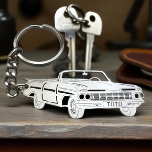 Può includere: Un portachiavi in metallo color argento con una sagoma ritagliata dettagliata di un'auto decappottabile classica. L'area della griglia anteriore mostra il testo TOTO. Il portachiavi include un anello diviso circolare fissato da una corta catena in metallo.