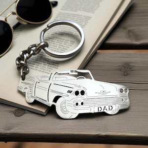 Può includere: Un portachiavi in metallo color argento con un design ritagliato di un'auto decappottabile d'epoca. La griglia anteriore dell'auto mostra la parola DAD in lettere maiuscole. Il portachiavi include un anello circolare standard collegato da una catena.