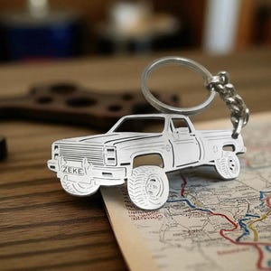 May include: A silver-coloured metal keyring shaped like a pickup truck featuring a detailed grille, wheels, and a number plate reading ZEKE. The truck silhouette is attached to a circular split ring and a short metal chain for holding keys securely.