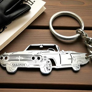 Può includere: Un portachiavi in metallo color argento con la sagoma ritagliata dettagliata di un'auto decappottabile d'epoca. Il design dell'auto include linee incise per i fari, la griglia e la carrozzeria, con la targa che mostra chiaramente 5XAD608.
