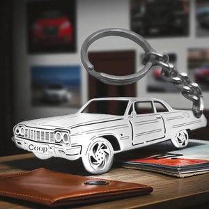 Könnte beinhalten: Ein silberfarbener Metallschlüsselanhänger in Form eines klassischen Autos mit detailliertem Kühlergrill, Scheinwerfern und Rädern. Das Wort Coop ist auf die vordere Stoßstange geprägt. Ein stabiler Metallring ist mit einer kurzen Kette am Dach befestigt.