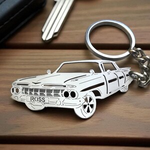 Può includere: Un portachiavi in metallo color argento a forma di auto d'epoca con griglia anteriore, fari e ruote dettagliati. La targa sul paraurti anteriore riporta la scritta ROSS in lettere nere. Il portachiavi è dotato di un anello circolare e catena.