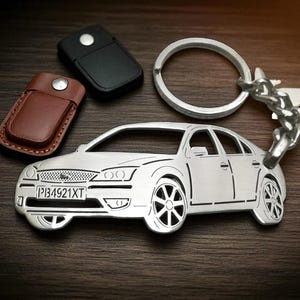 May include: A silver-colored metal keychain shaped like a sedan car with a license plate reading PB4921XT, attached to a circular metal ring. Next to it are two small rectangular leather-like key covers, one in black and one in brown, with silver snaps.