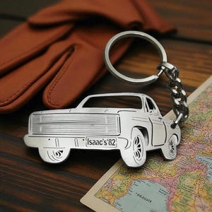May include: A silver-toned metal keyring shaped like a pickup truck with a detailed front grille and wheels. The number plate on the truck is engraved with the text Isaac's '82. The keyring includes a standard circular split ring and a short chain.