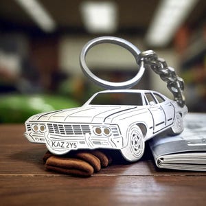 Op de afbeelding: Een zilverkleurige metalen sleutelhanger in de vorm van een klassieke sedan met zwarte gegraveerde omlijningen. De auto heeft een voorste nummerplaat met de tekst KAZ 2Y5 duidelijk zichtbaar. Een ronde ring is aan de bovenkant bevestigd.