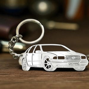 Può includere: Un portachiavi in metallo argentato con la sagoma ritagliata di una berlina. Il design presenta linee incise per i finestrini, le ruote e la griglia, con la targa che mostra chiaramente il testo GBIDYKNG in caratteri di colore nero.