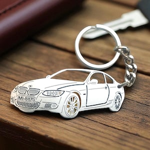 May include: A silver-toned metal keychain shaped like a BMW coupe. The car silhouette features detailed cutouts for the headlights, grille, and wheels. A license plate on the front bumper displays the text IMI-8870. It includes a circular ring.