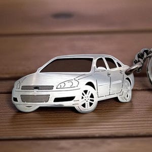 Op de afbeelding: Een zilverkleurige metalen sleutelhanger in de vorm van een sedan. De sleutelhanger heeft een geborstelde metalen afwerking met uitgesneden details die de ramen, wielen en grille van de auto voorstellen. Een metalen ring is aan de achterkant bevestigd.