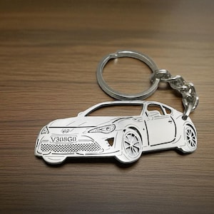 May include: A silver-colored metal keychain shaped like a sports car. The car silhouette features engraved details including headlights, wheels, a front grille, and a license plate with the text V308G0. A circular metal ring is attached by a chain.