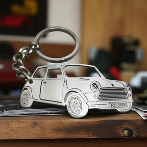 May include: A silver-toned metal keychain shaped like a classic Mini Cooper car. The keychain features detailed engravings of the car's body, wheels, and front grille, with the license plate text TRO 388W clearly visible on the front bumper.