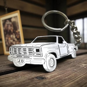 Könnte beinhalten: Ein silberfarbener Metallschlüsselanhänger in Form eines Ford F-150 Pick-up-Trucks. Der Schlüsselanhänger weist eingravierte schwarze Liniendetails auf, die die Karosserie, den Kühlergrill und die Räder des Trucks nachzeichnen. Ford steht auf dem Grill.