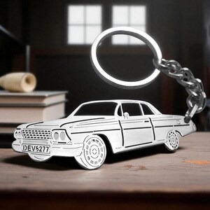 Può includere: Un portachiavi in metallo argentato a forma di auto classica con dettagli incisi in nero. L'auto presenta una griglia anteriore, ruote dettagliate e una targa che mostra chiaramente il testo DEV5277. È presente un anello metallico robusto.