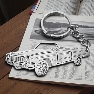 Könnte beinhalten: Ein silberfarbener Metallschlüsselanhänger in Form eines klassischen Cabriolets. Der Schlüsselanhänger zeigt die detaillierten Umrisse des Fahrzeugs, einschließlich Kühlergrill, Scheinwerfern und Rädern, und ist mit einer kurzen Kette an einem Ring befestigt.