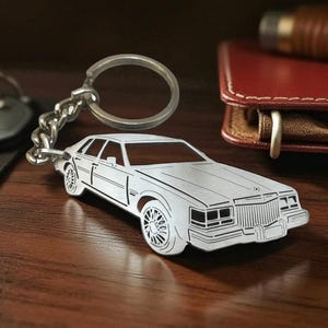 May include: A silver-toned metal keychain shaped like a classic sedan car. The keychain features a detailed outline of the vehicle, including the front grille, headlights, and wheels, attached to a circular split ring by a short metal chain.