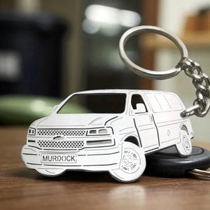 May include: A silver-coloured metal keyring shaped like a van with detailed cutouts of the front grille, headlights, and wheels. The front number plate area features the text MURDOCK engraved in black. A circular metal ring is attached to the top.
