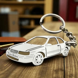 May include: A white metal keychain shaped like a two-door sedan car with black line details. The front bumper features the text "Toro Bravo" and a small Toyota logo. A silver-toned metal split ring is attached to the rear of the car silhouette.