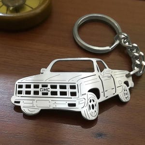 May include: A silver-toned metal keychain featuring a cutout silhouette of a GMC pickup truck. The design includes engraved lines detailing the grille, headlights, wheels, and body panels, attached to a circular split ring by a short metal chain.