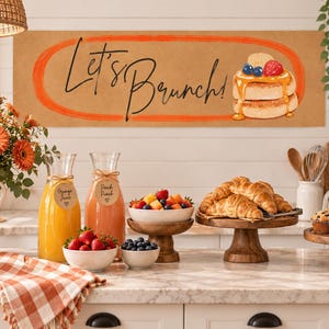 May include: A rectangular sign with a tan background featuring an orange oval border and the black cursive text Let's Brunch! next to an illustration of a stack of pancakes topped with syrup, blueberries, and a strawberry, displayed in a kitchen.