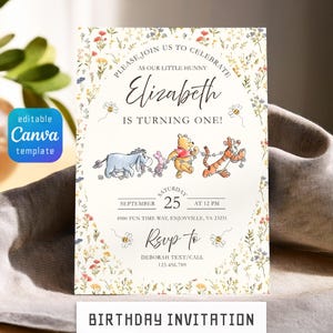 May include: A rectangular birthday invitation card featuring a floral border with Winnie the Pooh characters Eeyore, Piglet, Pooh, and Tigger. The text reads: PLEASE JOIN US TO CELEBRATE AS OUR LITTLE HUNNY ELIZABETH IS TURNING ONE! SEPTEMBER 25 AT 12 PM.
