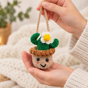 May include: A small, crocheted cactus ornament featuring a green body with a white and yellow flower, sitting in a tan pot with a smiling face, pink cheeks, and black eyes. The cactus is attached to a braided tan cord for hanging purposes.