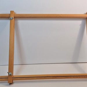 May include: A rectangular wooden embroidery frame consisting of four adjustable light-colored wooden bars. The frame is held together at each of the four corners by metal wing nuts and bolts, allowing for the adjustment of the frame's dimensions.