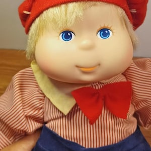 May include: A doll with blonde hair, blue eyes, and a soft face wearing a red beret, a red and white striped long-sleeve shirt with a yellow collar, a red bow tie, and dark blue pants. The doll has a rounded head and a gentle, smiling expression.